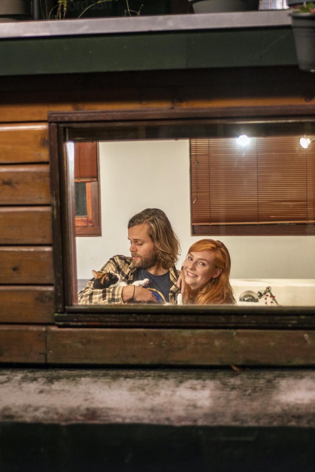 Door het raam zie je een jong koppel in hun woonboot staan, een kat in hun armen