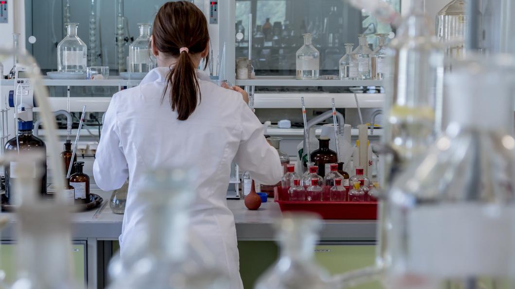 Een onderzoeker staat in een labratorium tussen de erlemeyers en ander onderzoeksmateriaal