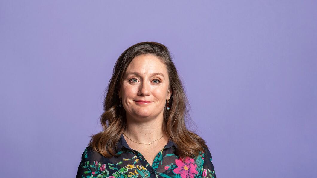 Foto van Suzanne Kröger, een witte vrouw. Ze draagt een zwarte blouse met groene en rode planten erop, heeft bruin haar en heeft een kleine glimlach.