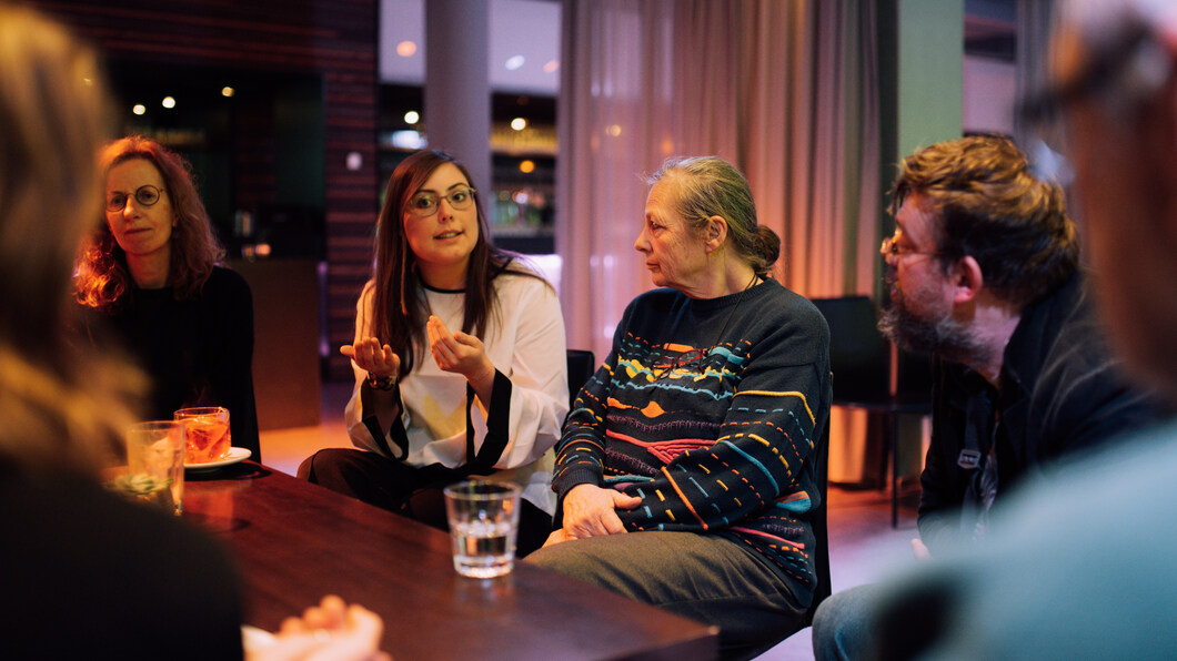 Een jong persoon zit aan tafel, pratend met veel handgebaren. Een ouder persoon luistert. Om hen heen zitten nog meer luisterende mensen.