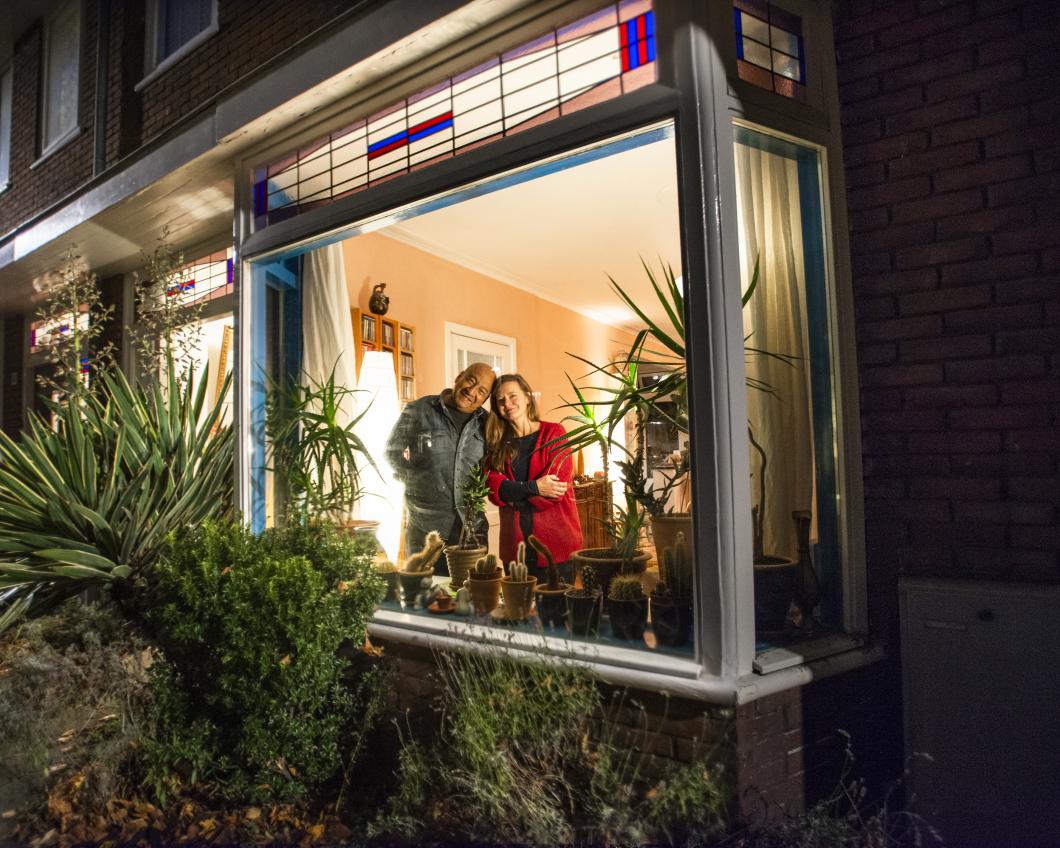 Een koppel staat samen achter hun raam, het is donker buiten en binnen brand warm licht