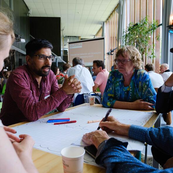 Vijf mensen zitten rond een tafel. Een man van kleur met donker haar en een rode blouse spreekt. De vier anderen, een witte vrouw van middelbare leeftijd, twee witte mannen met een pet op, en een jonge vrouw met blond haar luisteren aandachtig.